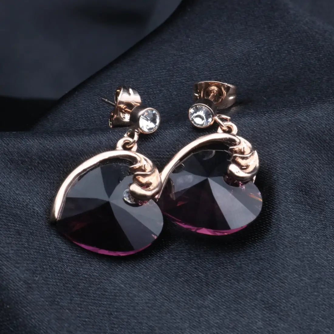 A pair of gold-plated stainless steel earrings with a heart-shaped design featuring a central American diamond, displayed against a black fabric background.