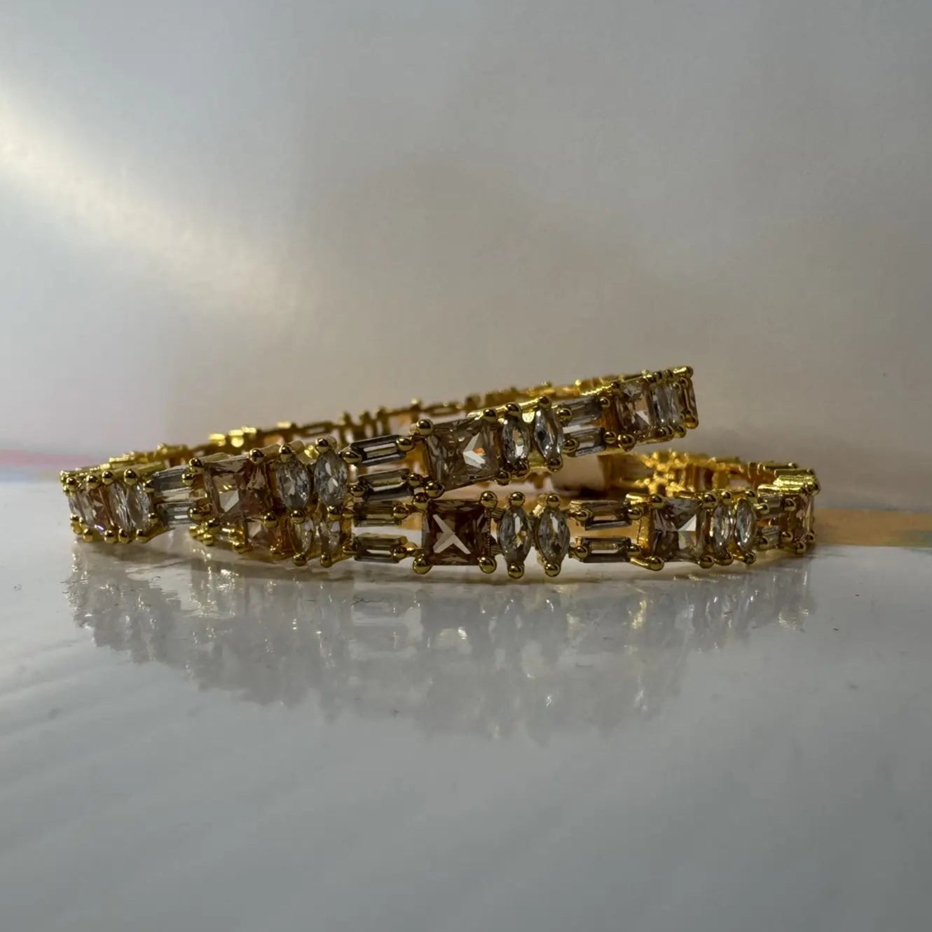 Gold plated bangles with clear gemstones in different patterns on a reflective surface with white background