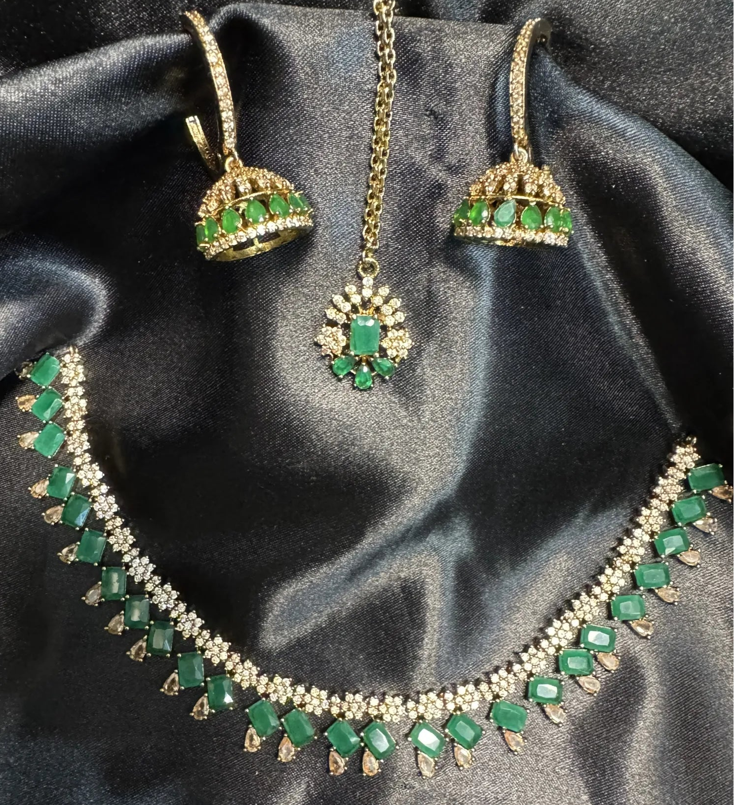 JV52 necklace set featuring emerald green CZ stones, gold-plated jhumki earrings, and a teeka displayed on black satin.