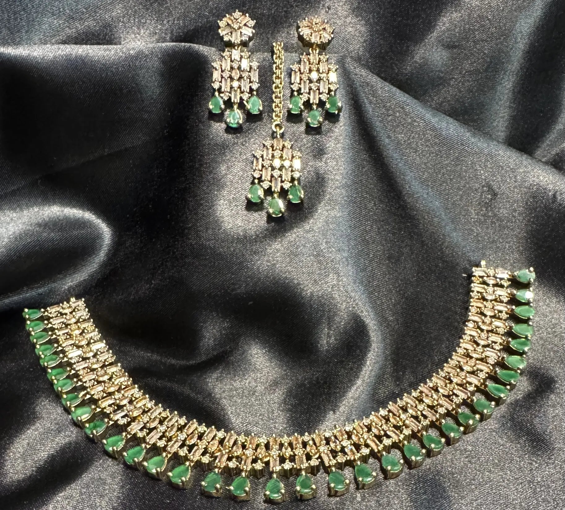 JV53 emerald necklace set with baguette CZ stones, matching earrings, and teeka displayed on a black satin background.