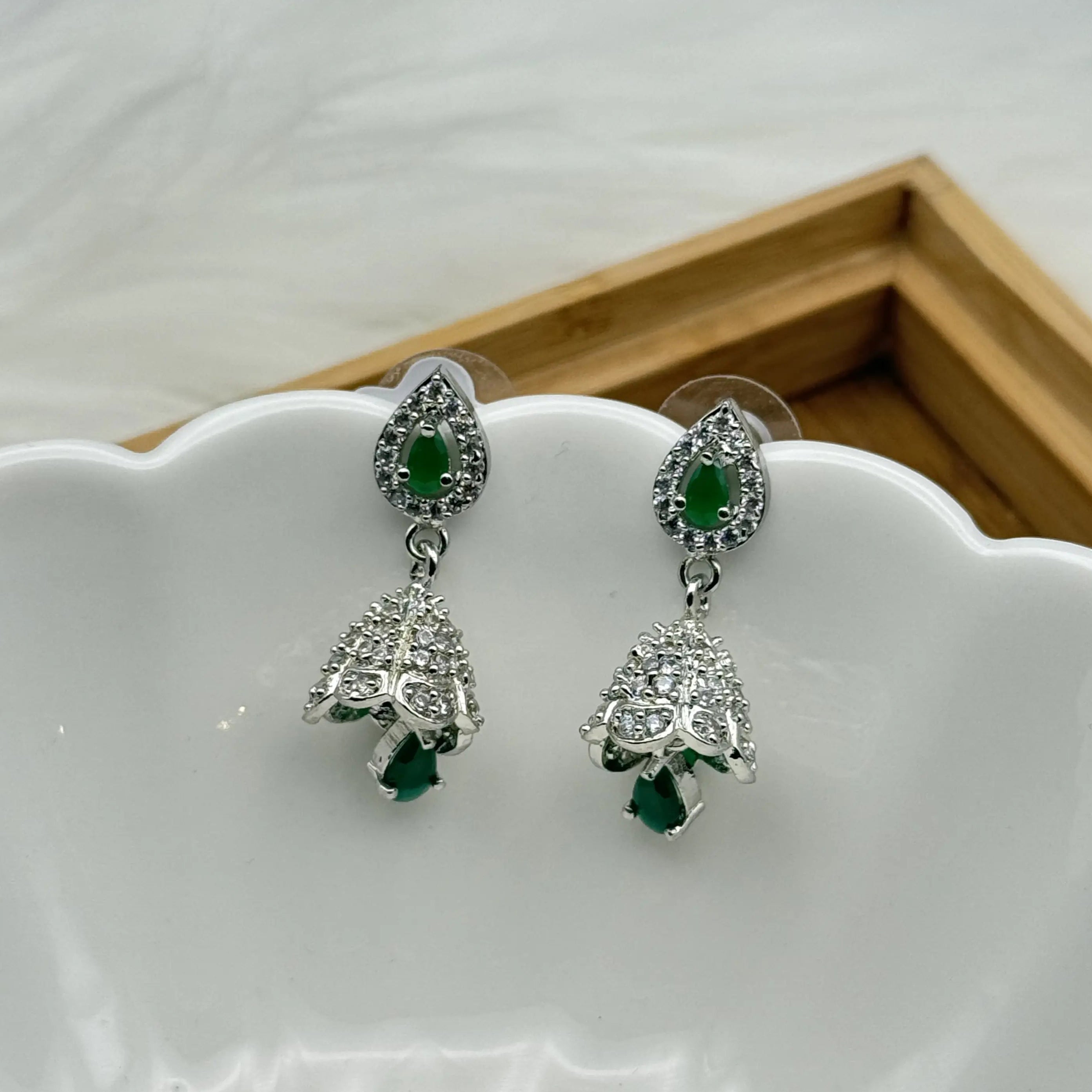 Pair of silver Pakistani style jhumka earrings with green gemstones and a hanging bell like design on a white dish.