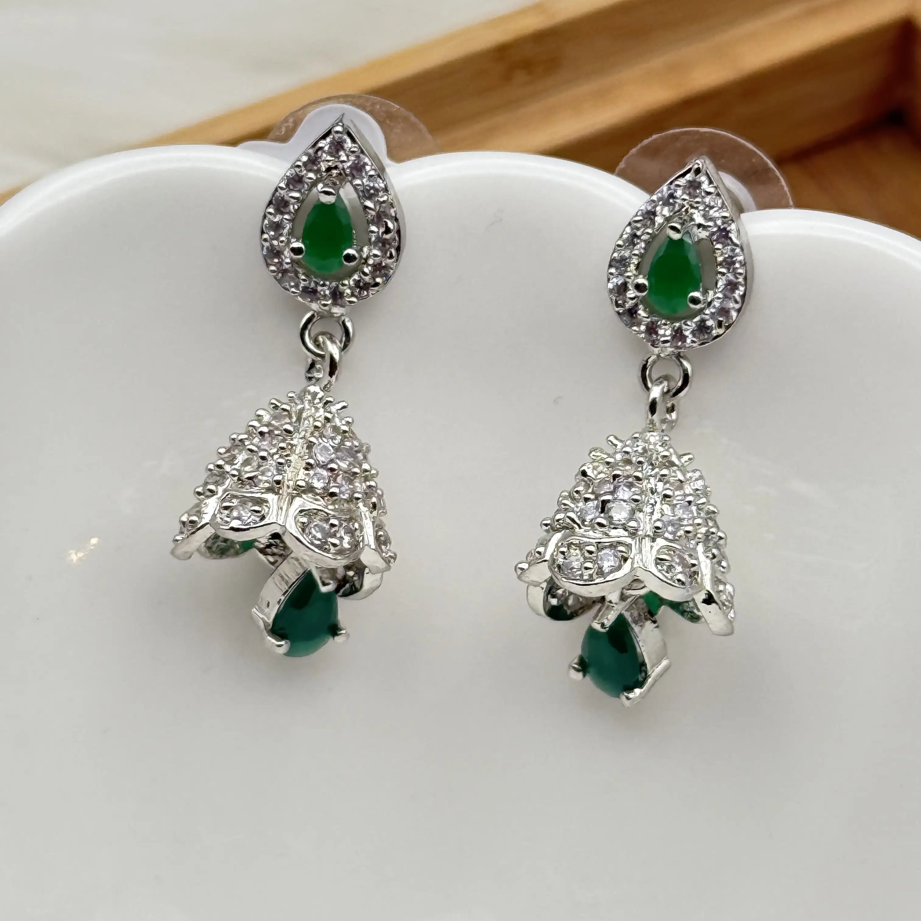 another angle of pair of silver Pakistani style jhumka earrings with green gemstones and a hanging bell like design on a white dish.