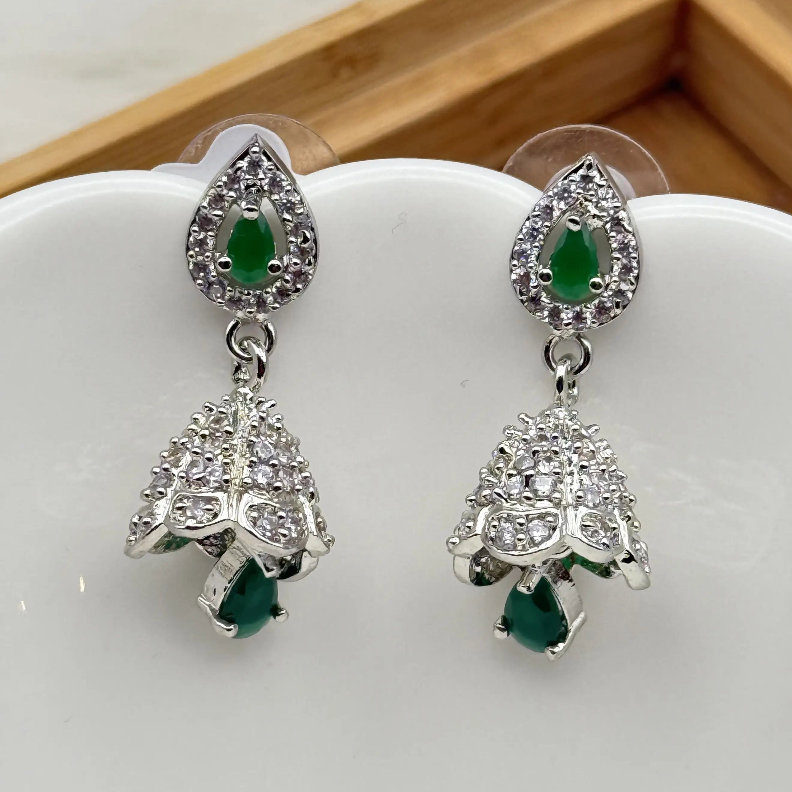 top view of Pair of silver Pakistani style jhumka earrings with green gemstones and a hanging bell like design on a white dish.