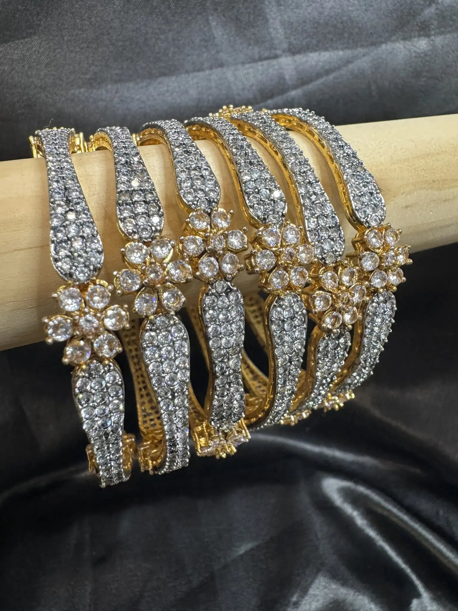 Set of gold-plated, American diamond bangles with flower patterns and clear gemstones on a wooden stick and black silk background