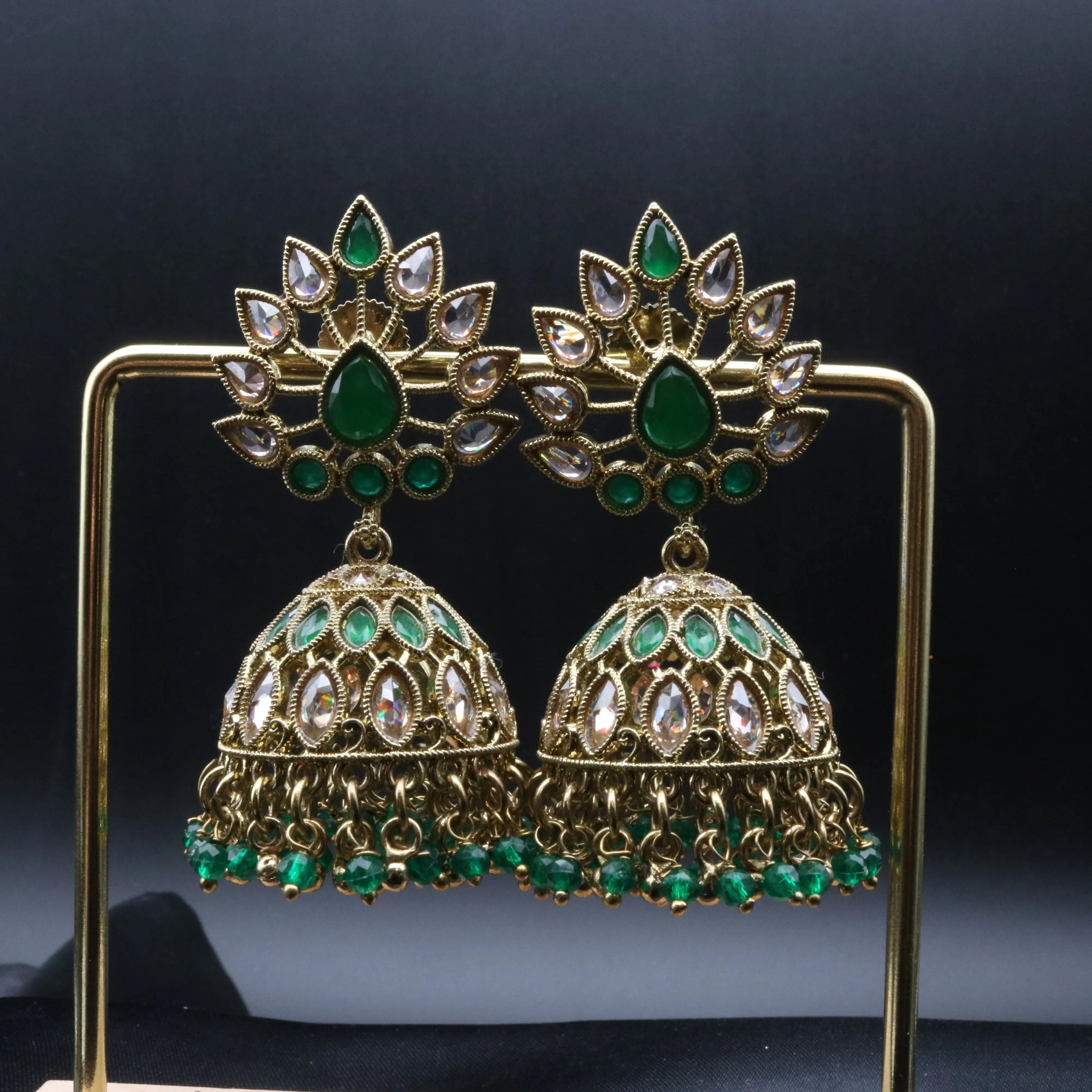 Pakistani style jhumka earrings with gold base and green and clear gemstones on a stand against a dark background