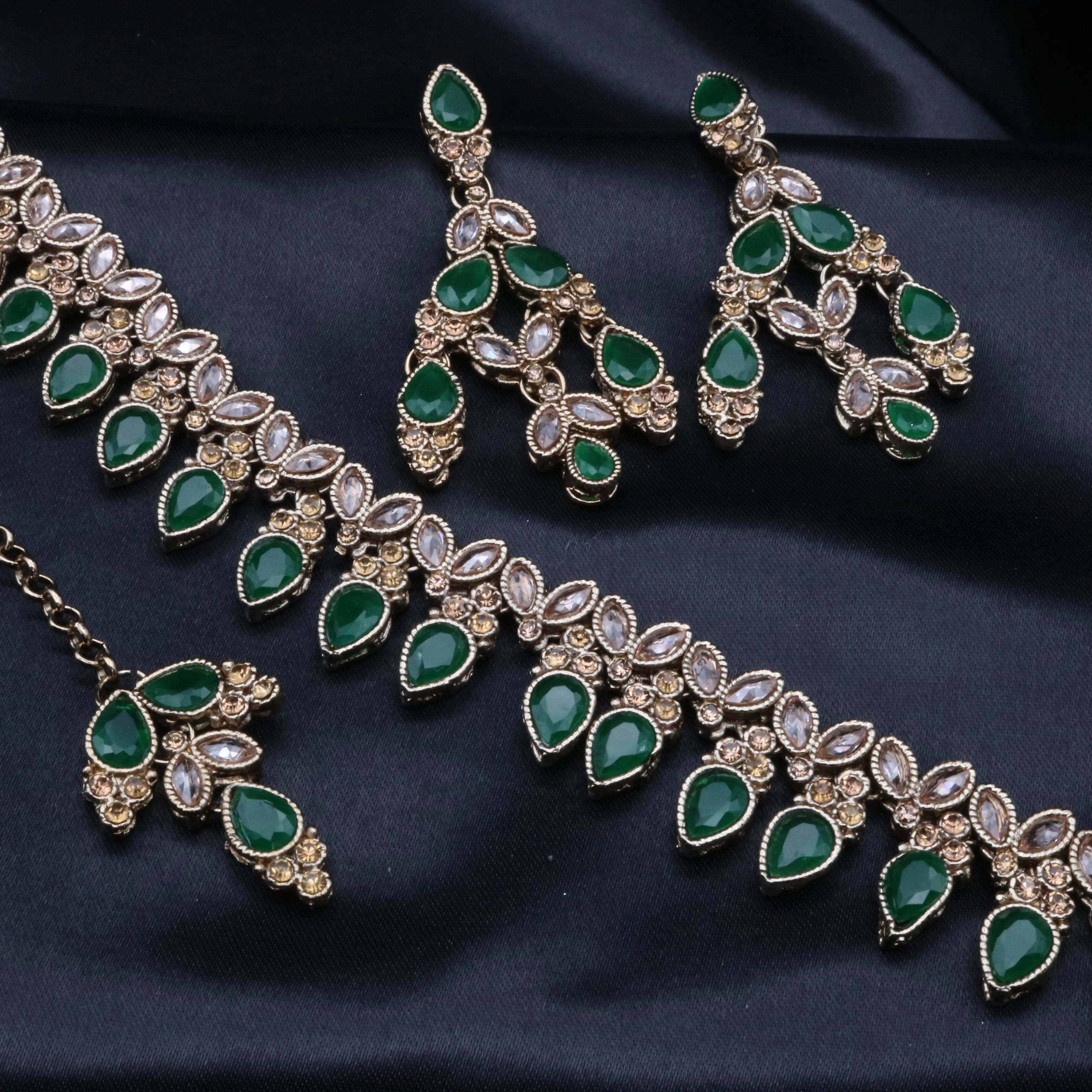 Cleo necklace set in emerald green featuring pear-cut stones and floral CZ details on gold base, laid on black fabric.