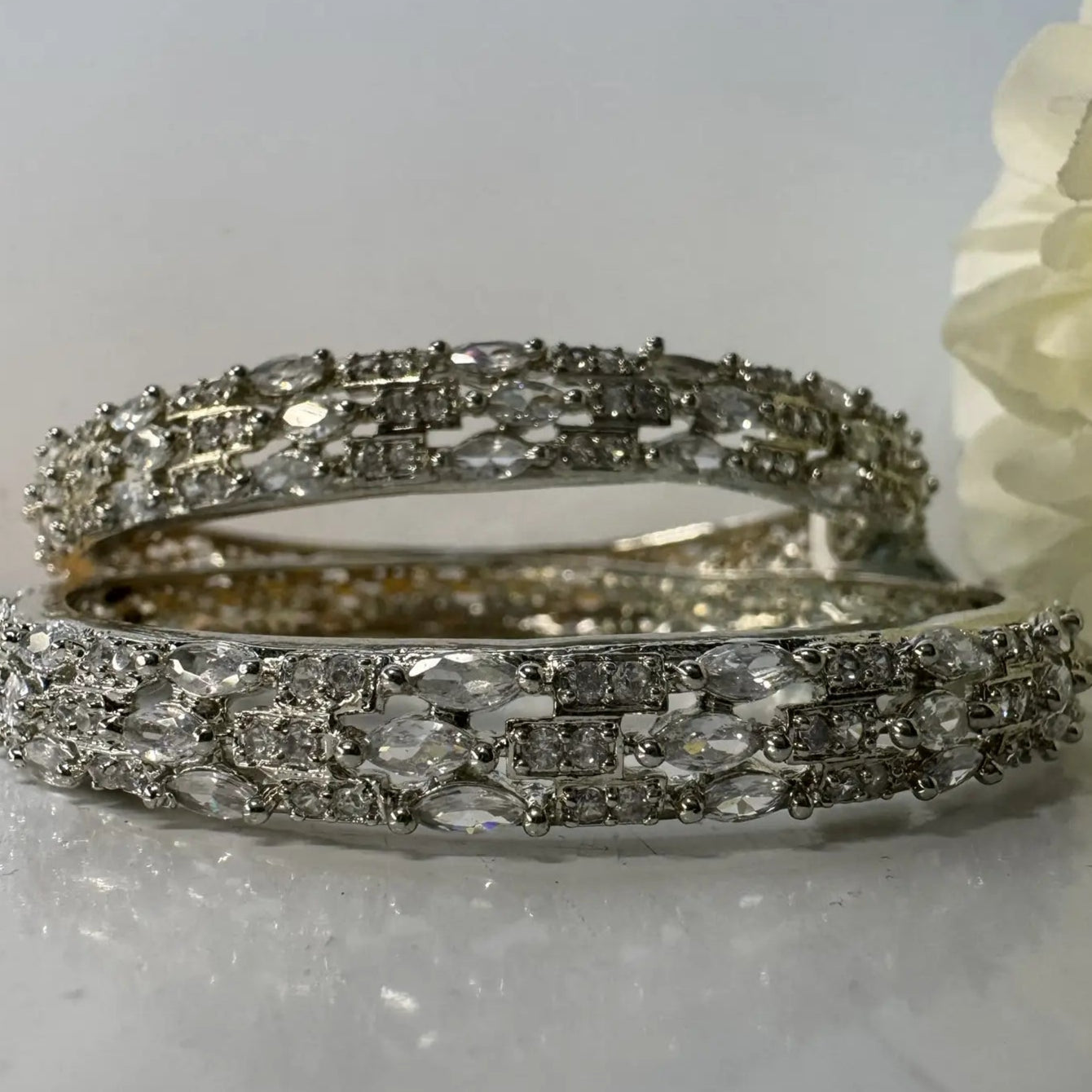 American diamonds bangles in silver with clear gemstones on a white surface and white flower at the back