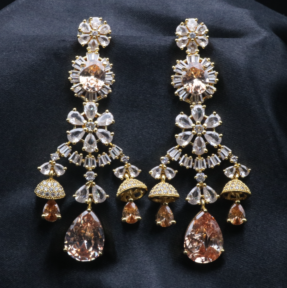 A pair of stainless steel and gold-plated statement earrings with intricate designs and multiple layers of sparkling stones displayed against a dark background.
