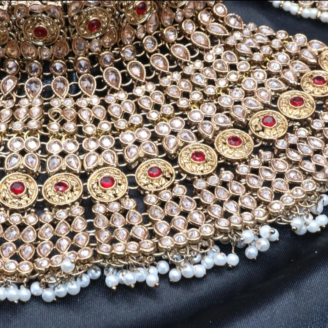 A gold-colored Pakistani style bridal set with intricate designs and pearl-like beads, displayed alongside a pair of matching jhumka earrings and matha patti on a dark fabric background.