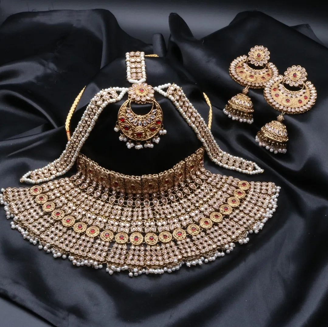 A gold-colored Pakistani style bridal set with intricate designs and pearl-like beads, displayed alongside a pair of matching jhumka earrings and matha patti on a dark fabric background.