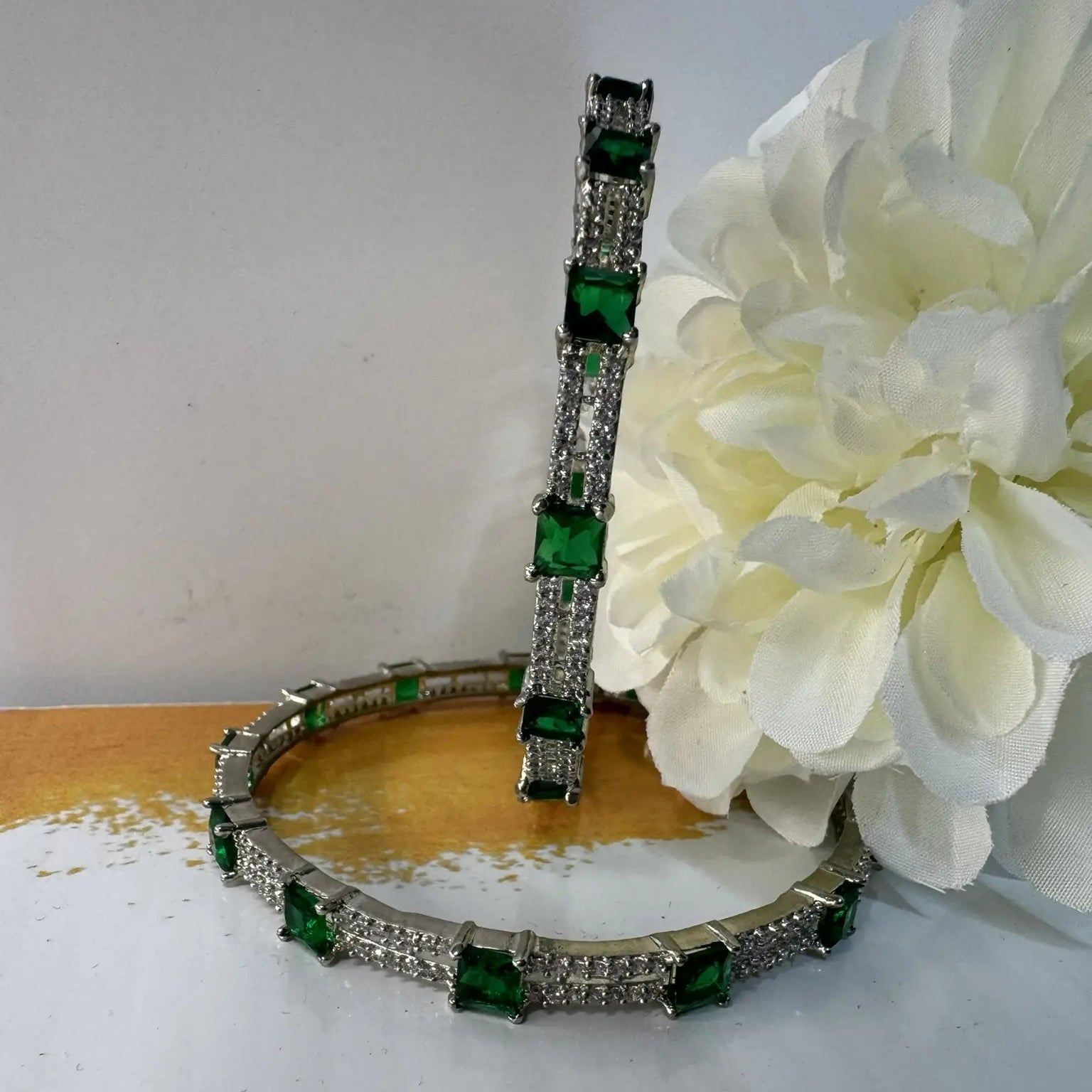 Silver bangles with green gemstones on a white surface with flowers