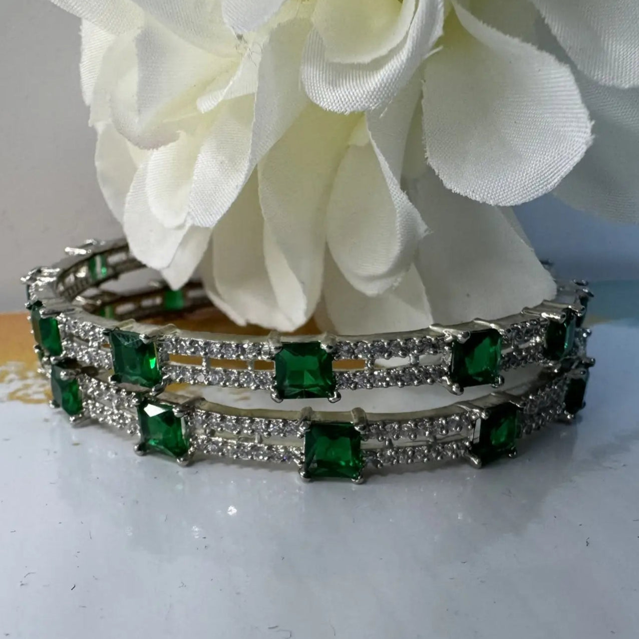 Bangles with green gemstones on a white surface with flowers in the background