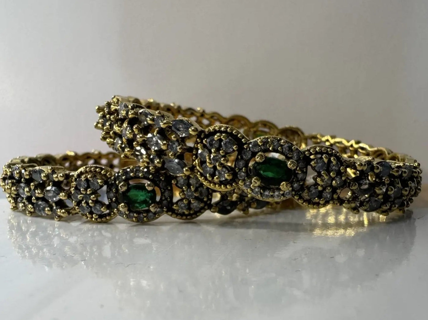 A set of gold-plated stainless steel bangles adorned with American diamond accents and green gemstones on a white surface