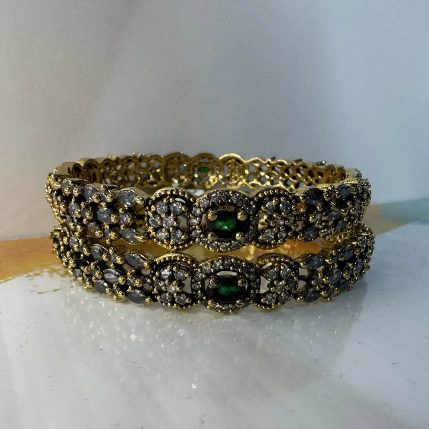 A set of gold-plated stainless steel bangles adorned with American diamond accents and green gemstones on a white colorful surface