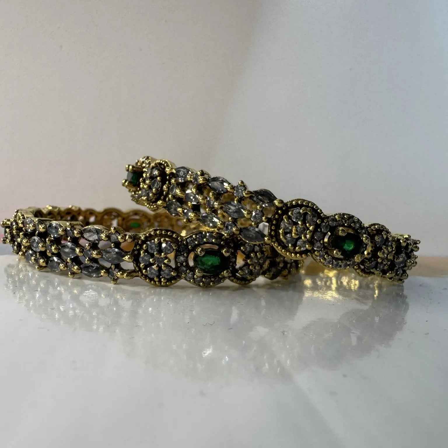 A set of gold-plated stainless steel bangles adorned with American diamond accents and green gemstones on a white background