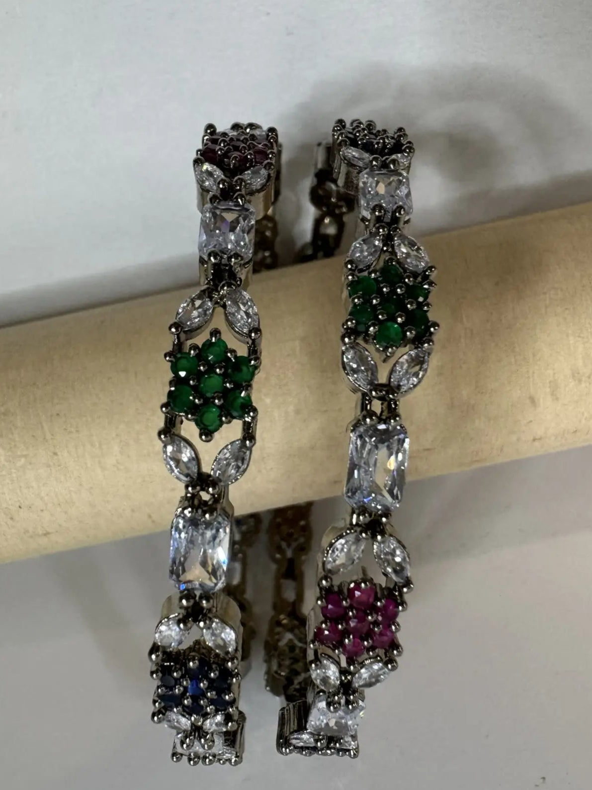 A multicolored diamond-encrusted bangles with vibrant hues of blue, green, and pink, set on a silver base, displayed against a textured white background on a beige stick against
