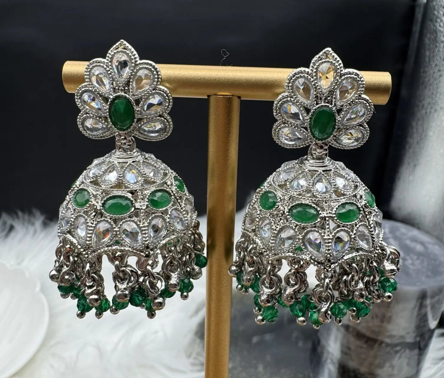 another angle of pair of Pakistani style silver jhumka earrings with green gemstones on a gold stand.