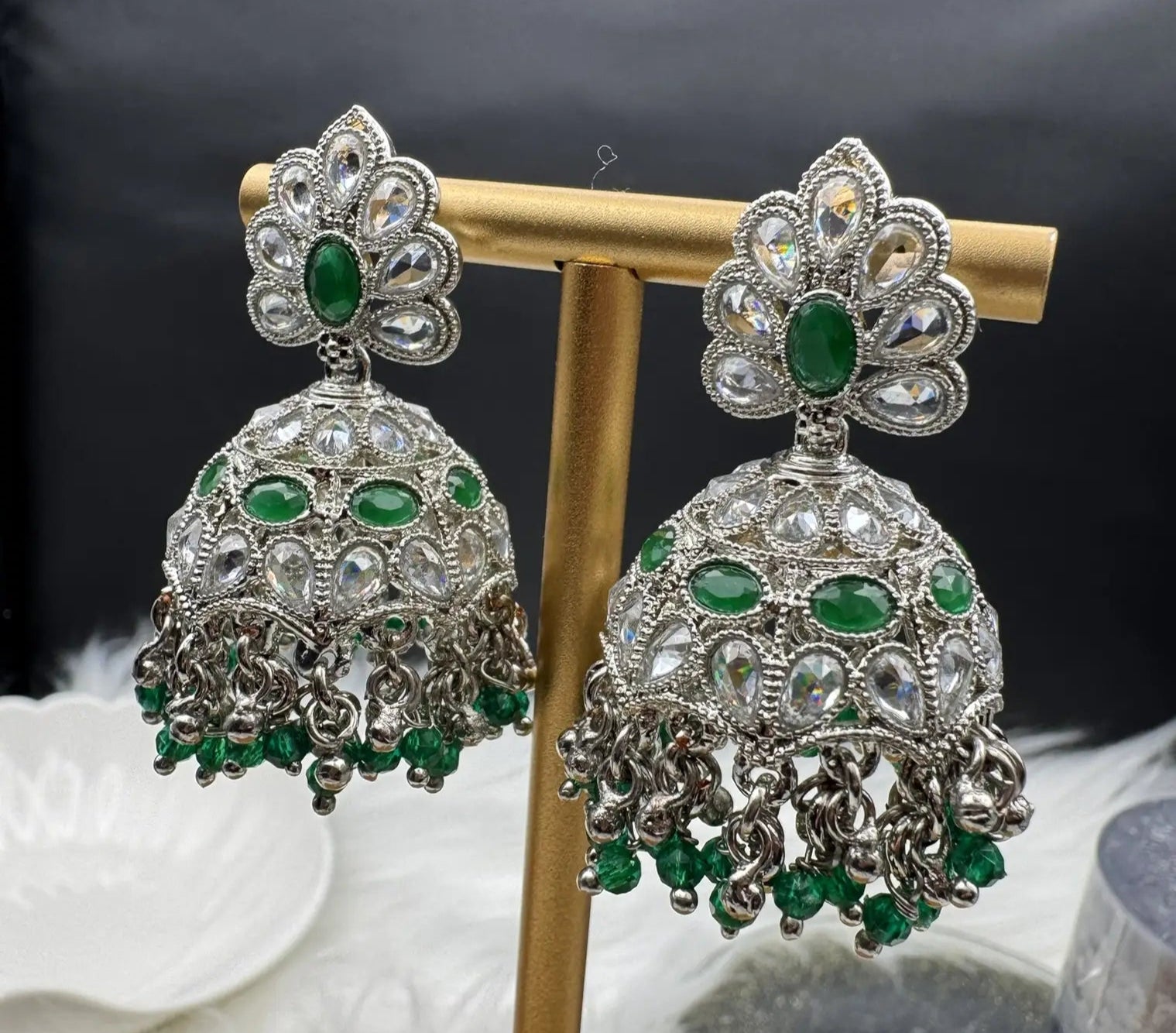 Ornate silver jhumka earrings with green gemstones on a stand against a dark background