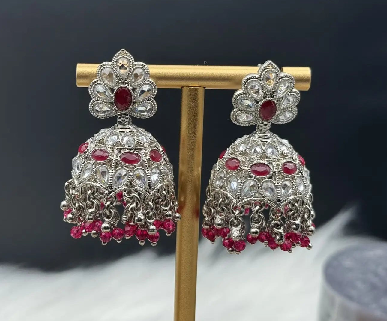 frontal view of pair of Pakistani style silver jhumka earrings with red gemstones on a gold stand.