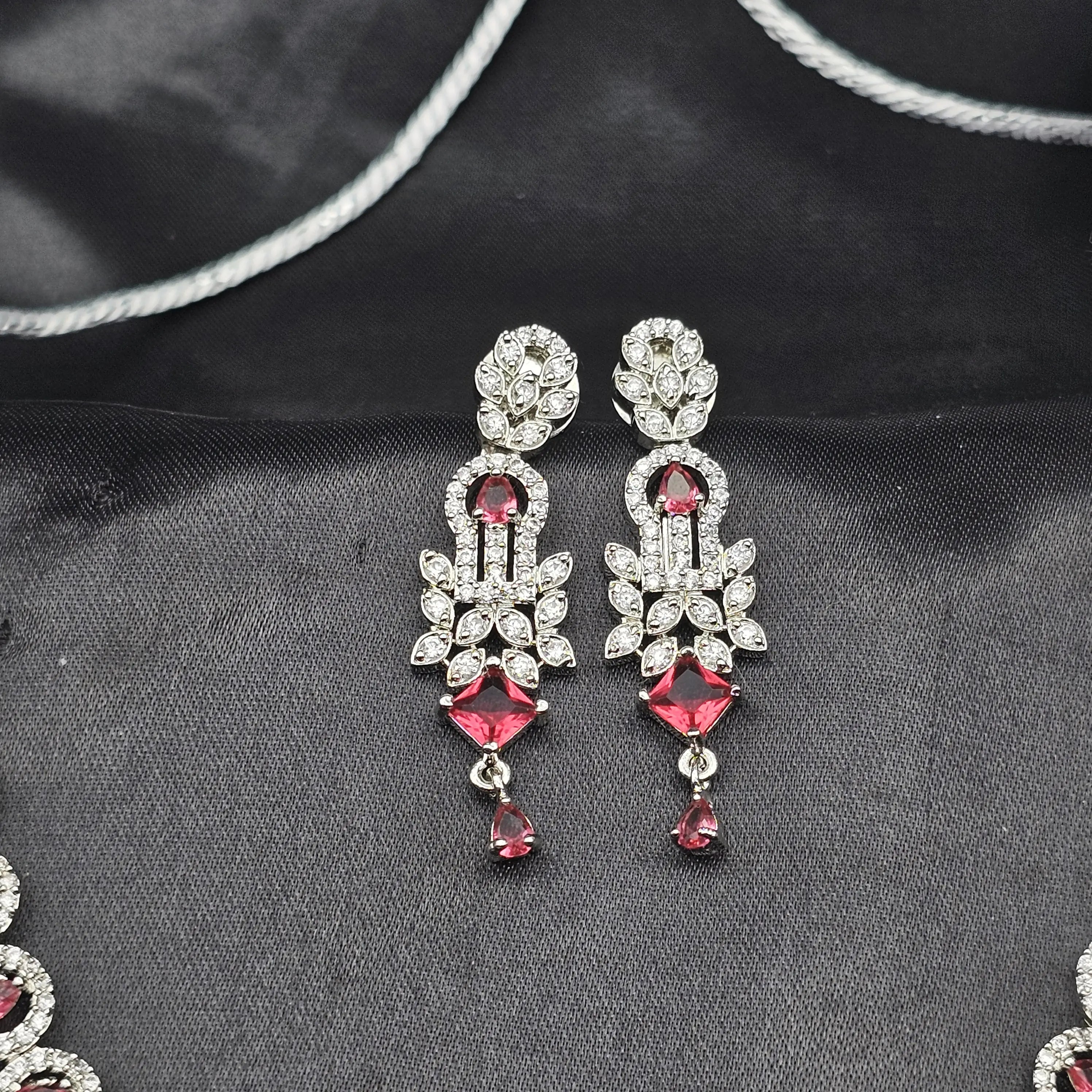 Euphoric earring pair with ruby CZ stones, floral CZ embellishments, and teardrop dangles on a black satin background.