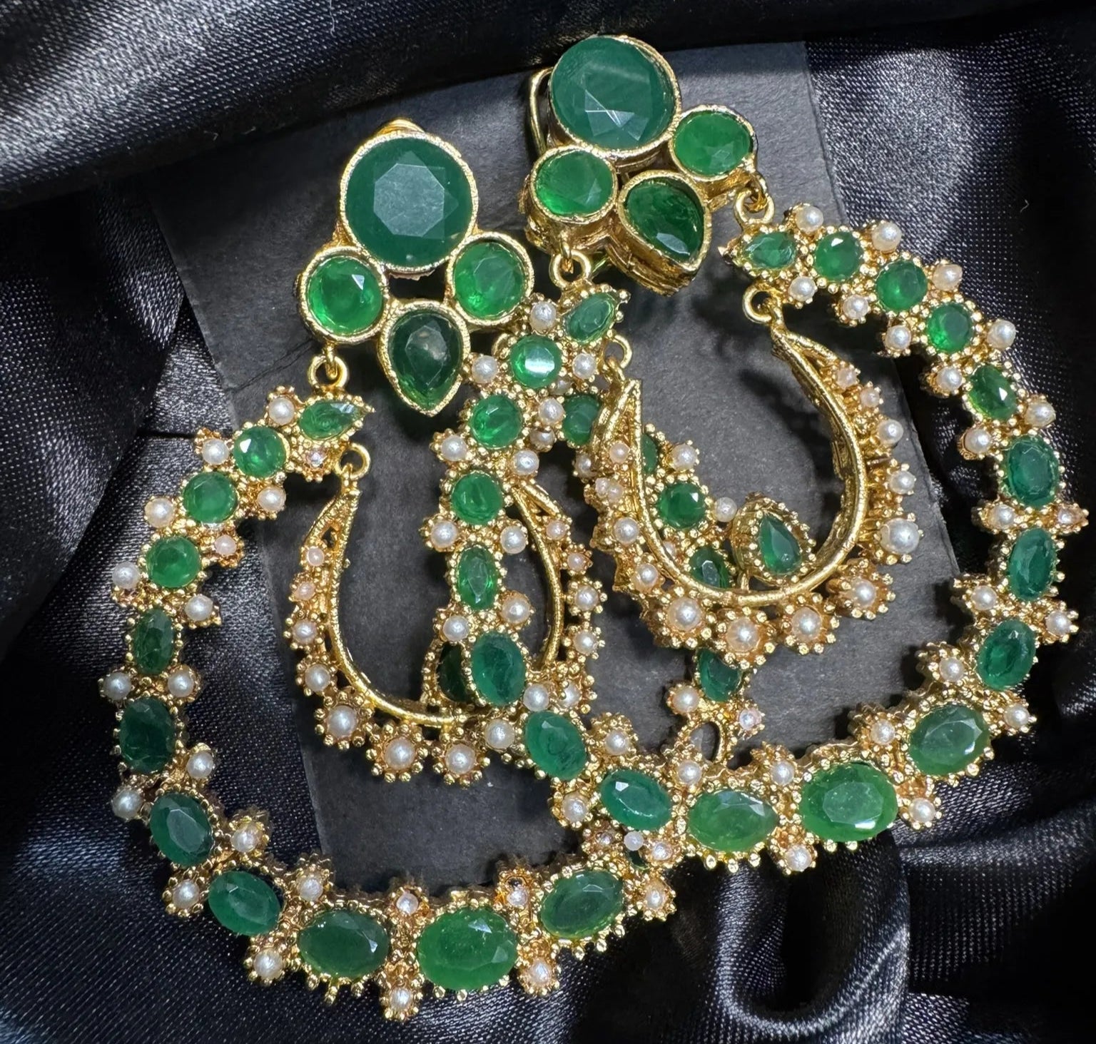 Ornate earrings with green gemstones and gold accents on a black satin background