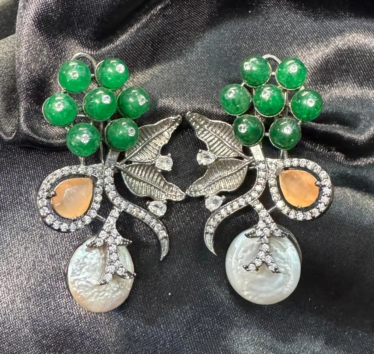 Pair of earrings with green gemstones and pearl-like elements on a dark fabric background