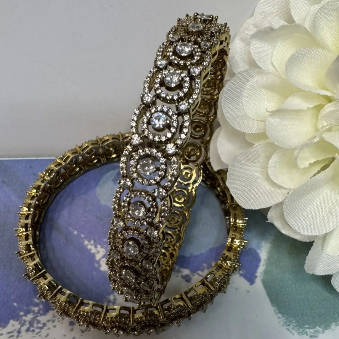 Gold bangles with intricate design on a white surface with a white flower