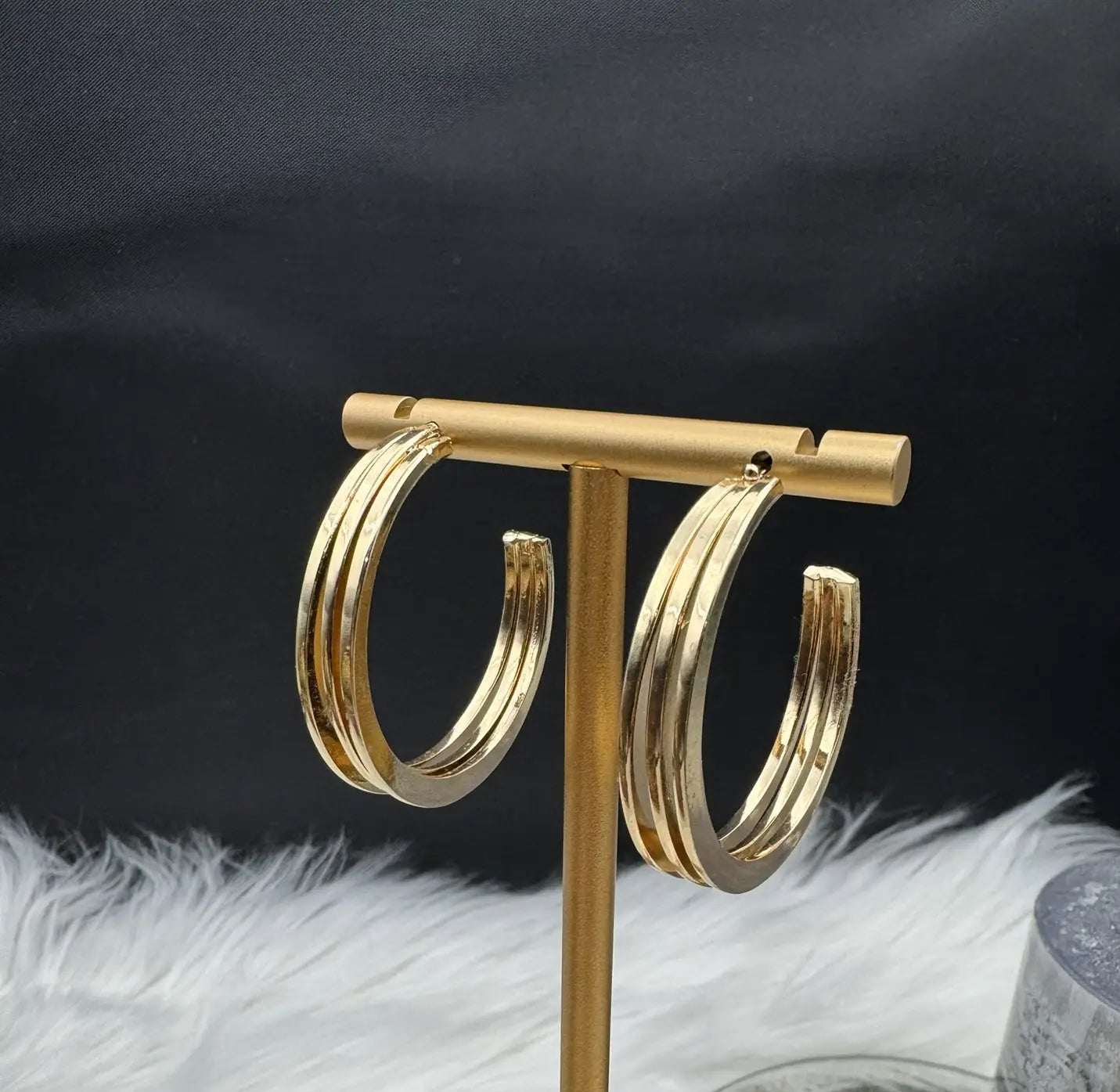 another angle of gold plated hoop earrings on a gold stand against a dark background with white feathers.