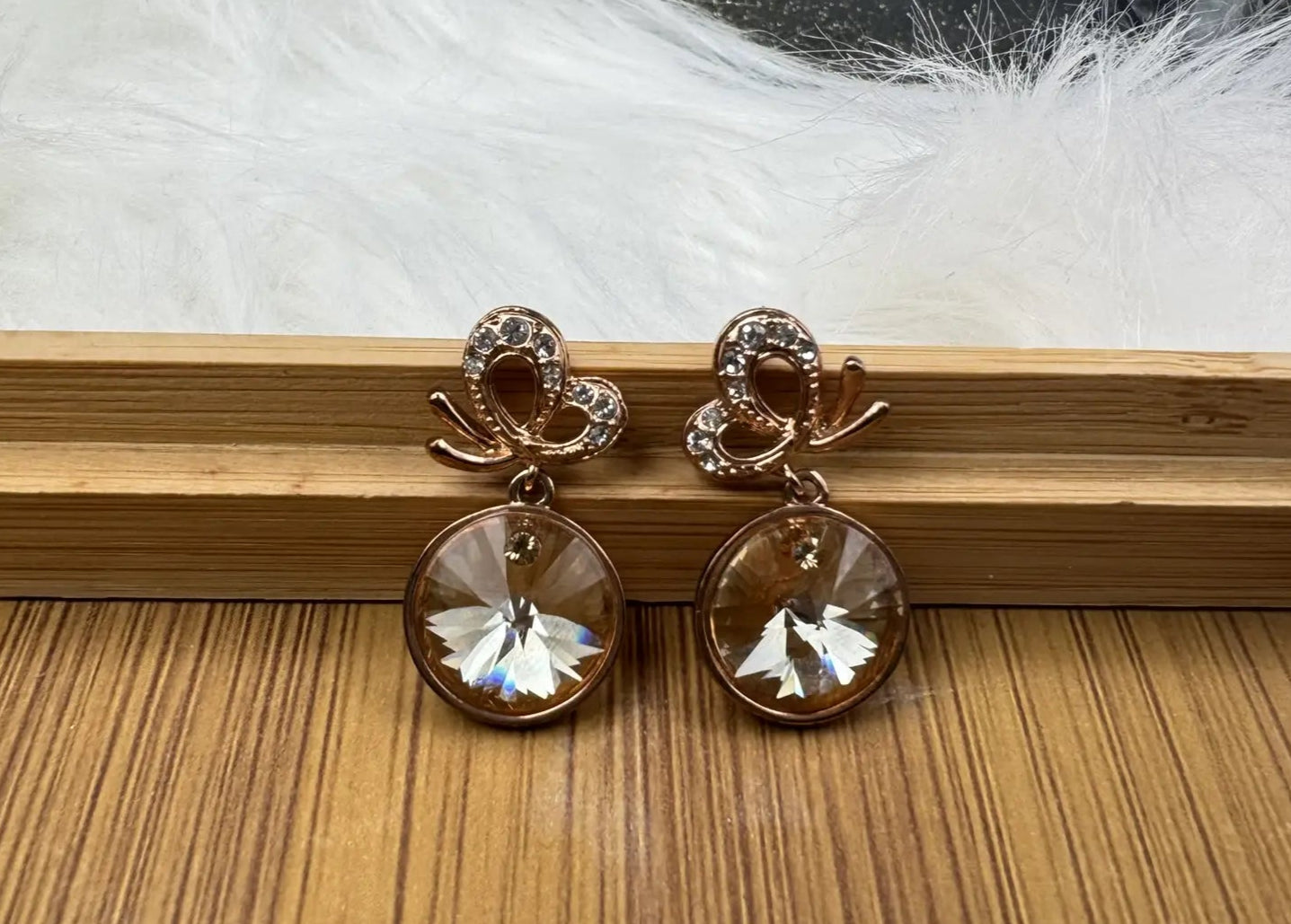 Pair of gold plated earrings with clear gemstones on a wooden surface with white feathers in the background