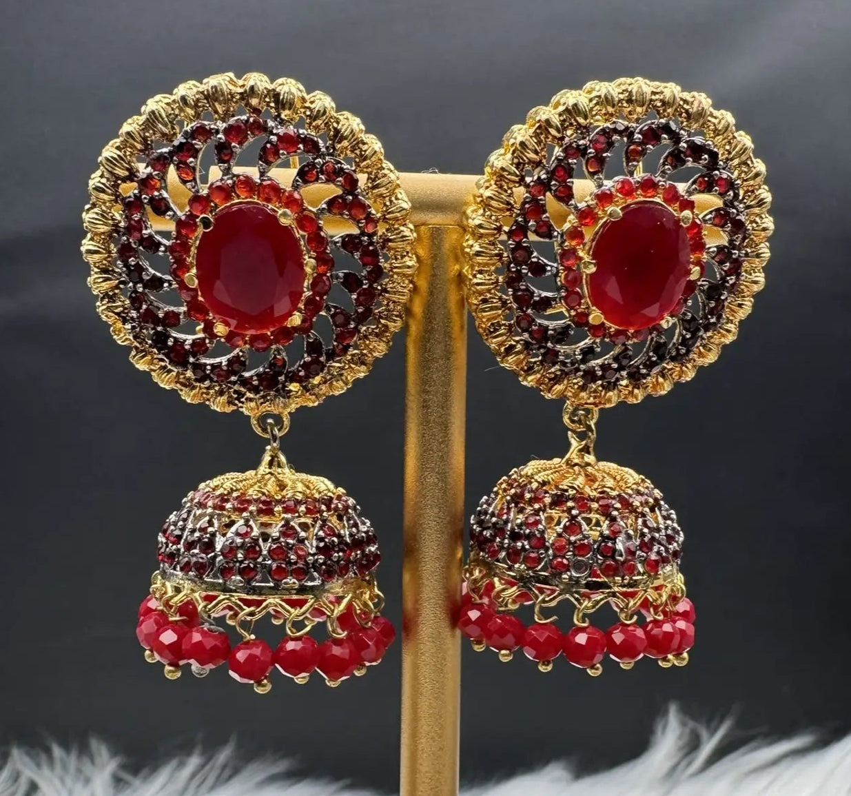 frontal view of Pakistani style jhumka earrings gold-plated and red colored gemstones on a stand with a dark background