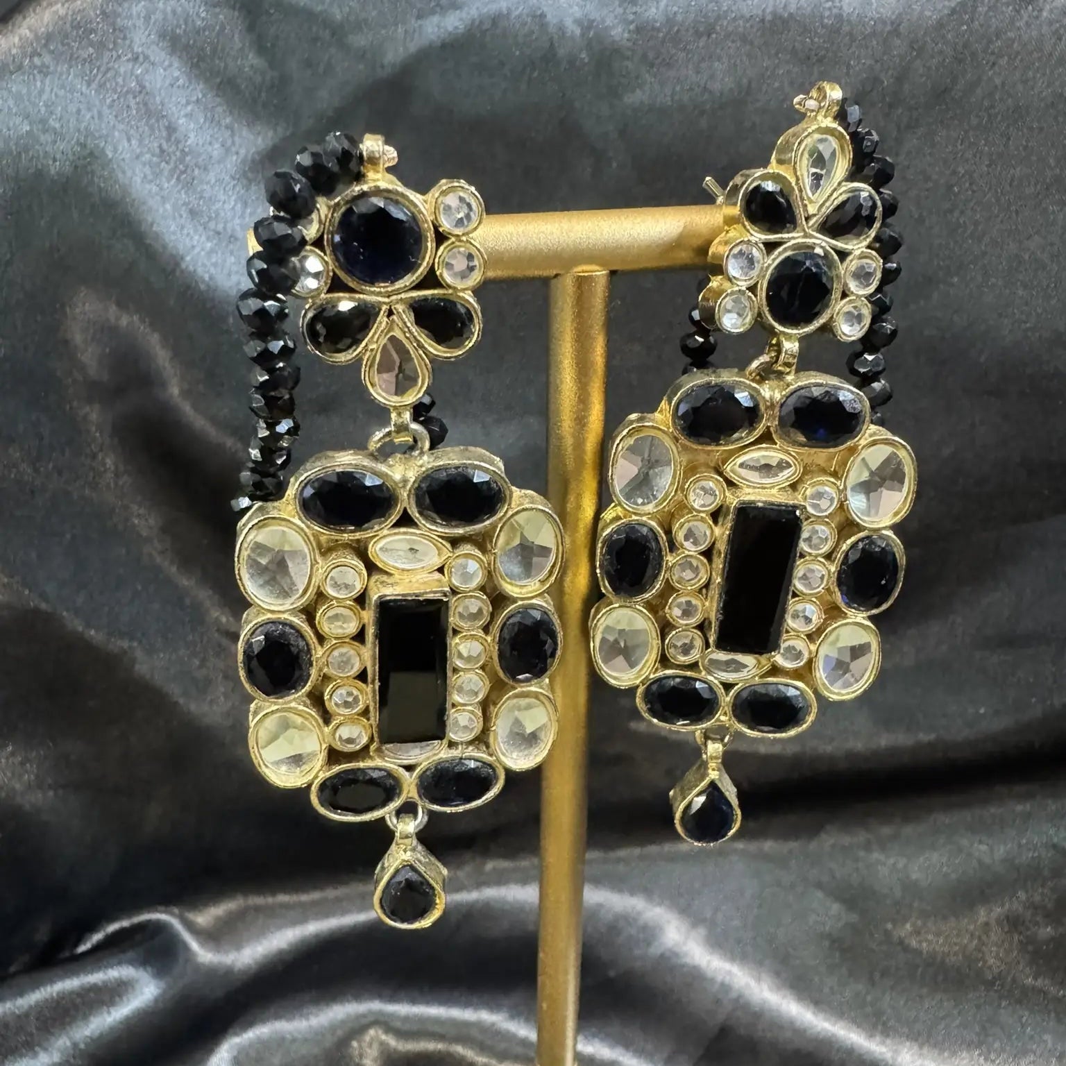 Ornate earrings with black and gold details on a stand against a dark fabric background