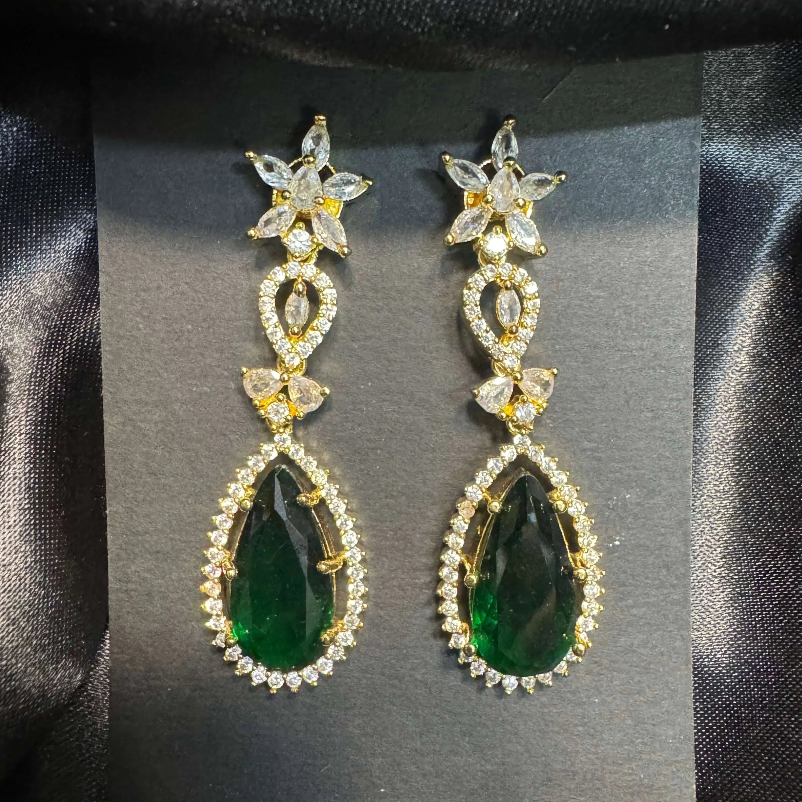 Pair of emerald teardrop earrings with gold accents and American diamonds on a black card.