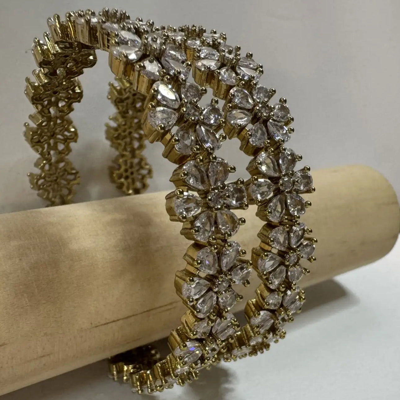 24k gold plated floral pattern bangle set with antique polish for a lasting shine with clear gemstones on a wooden stick against a white background