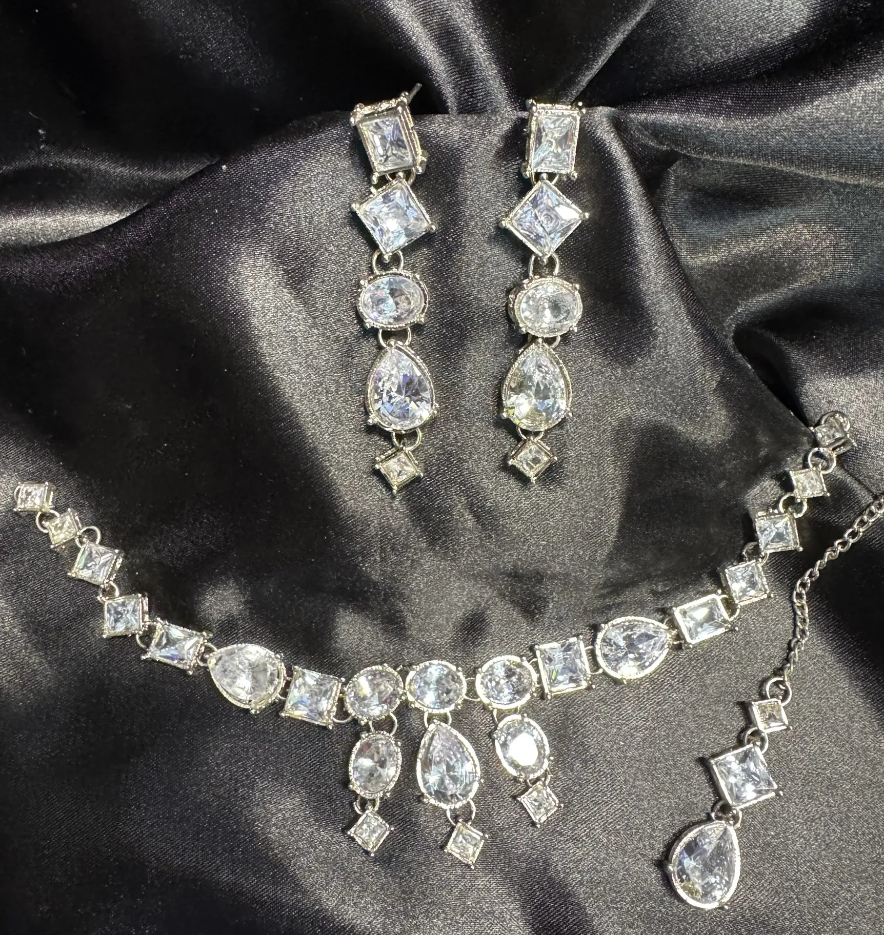 JV147 necklace set featuring clear cubic zirconia stones in square, round, and teardrop shapes laid on black satin fabric.