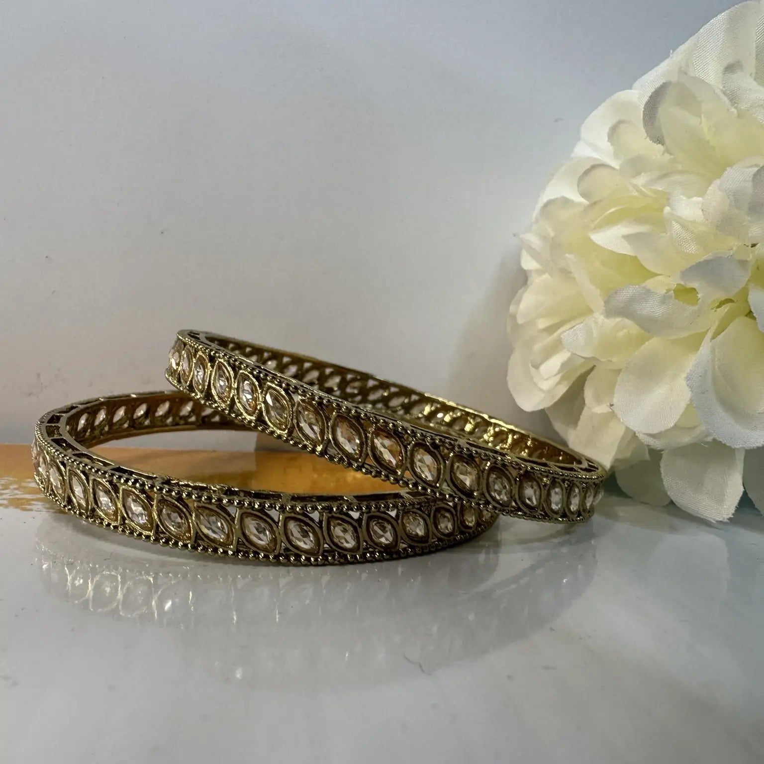 Two gold bangles with intricate designs on a white surface with a white flower in the background.