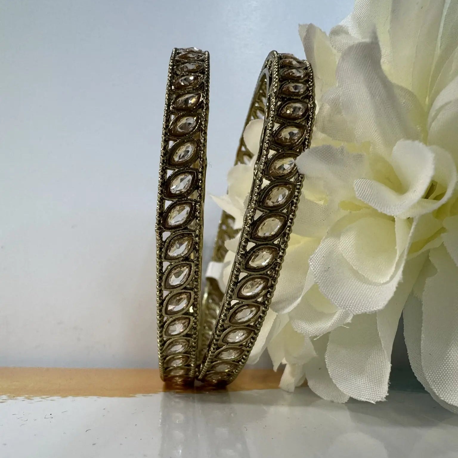 Decorative gold bangles with clear gemstones on a light surface with white flowers.