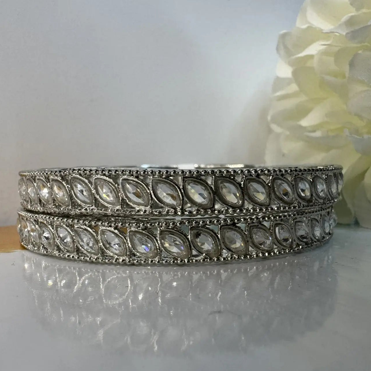Silver bangles with clear gemstones on a reflective surface with a light background