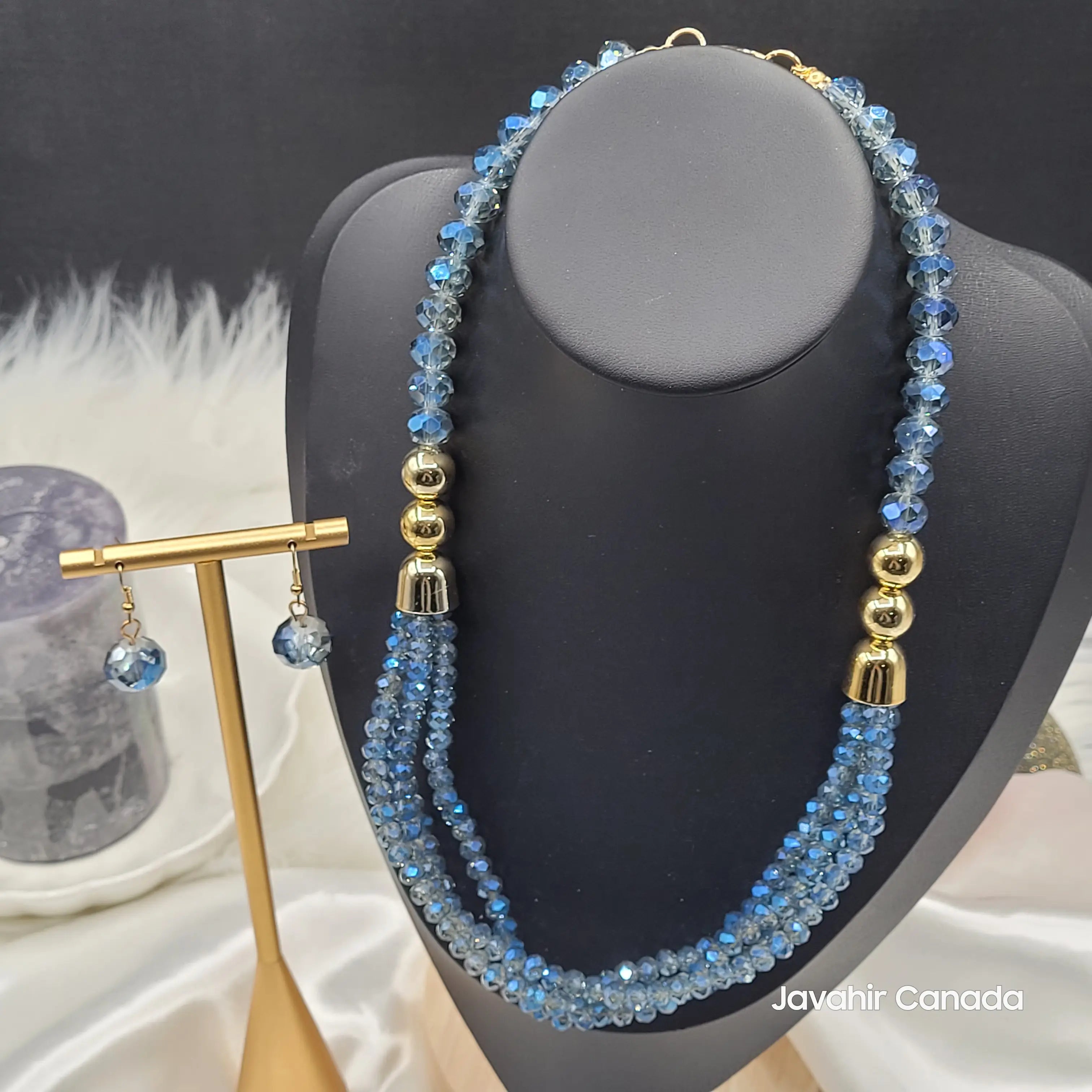 full view of the JV161 necklace set in vibrant sky blue. Multi-faceted crystal beads are arranged in three elegant strands with shiny gold-tone spacers. Includes matching drop earrings displayed on a stand.
