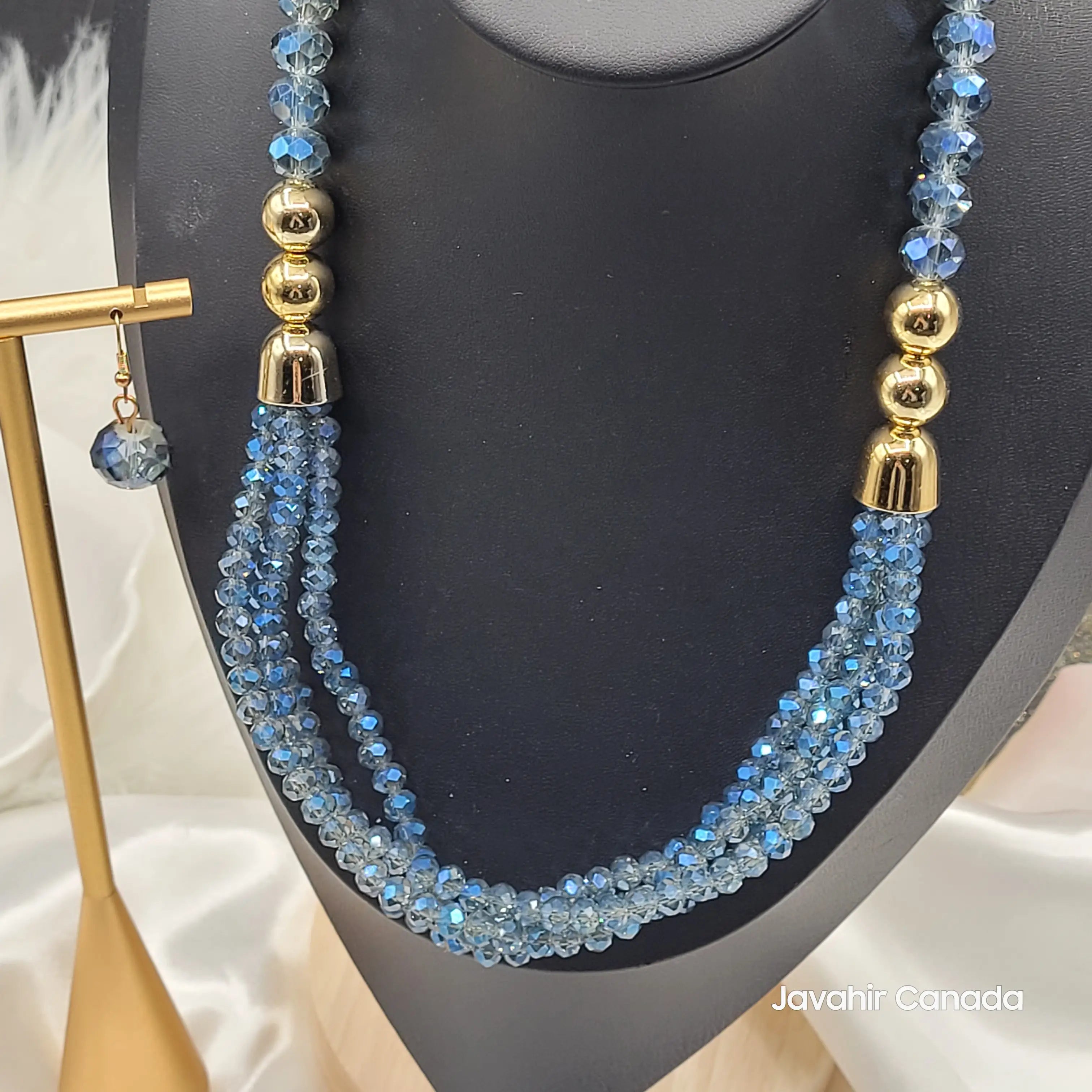Close-up view of the JV161 necklace in vibrant sky blue crystal beads, featuring a three-strand twisted design and shiny gold-toned accents. One matching earring is also visible on a display stand.