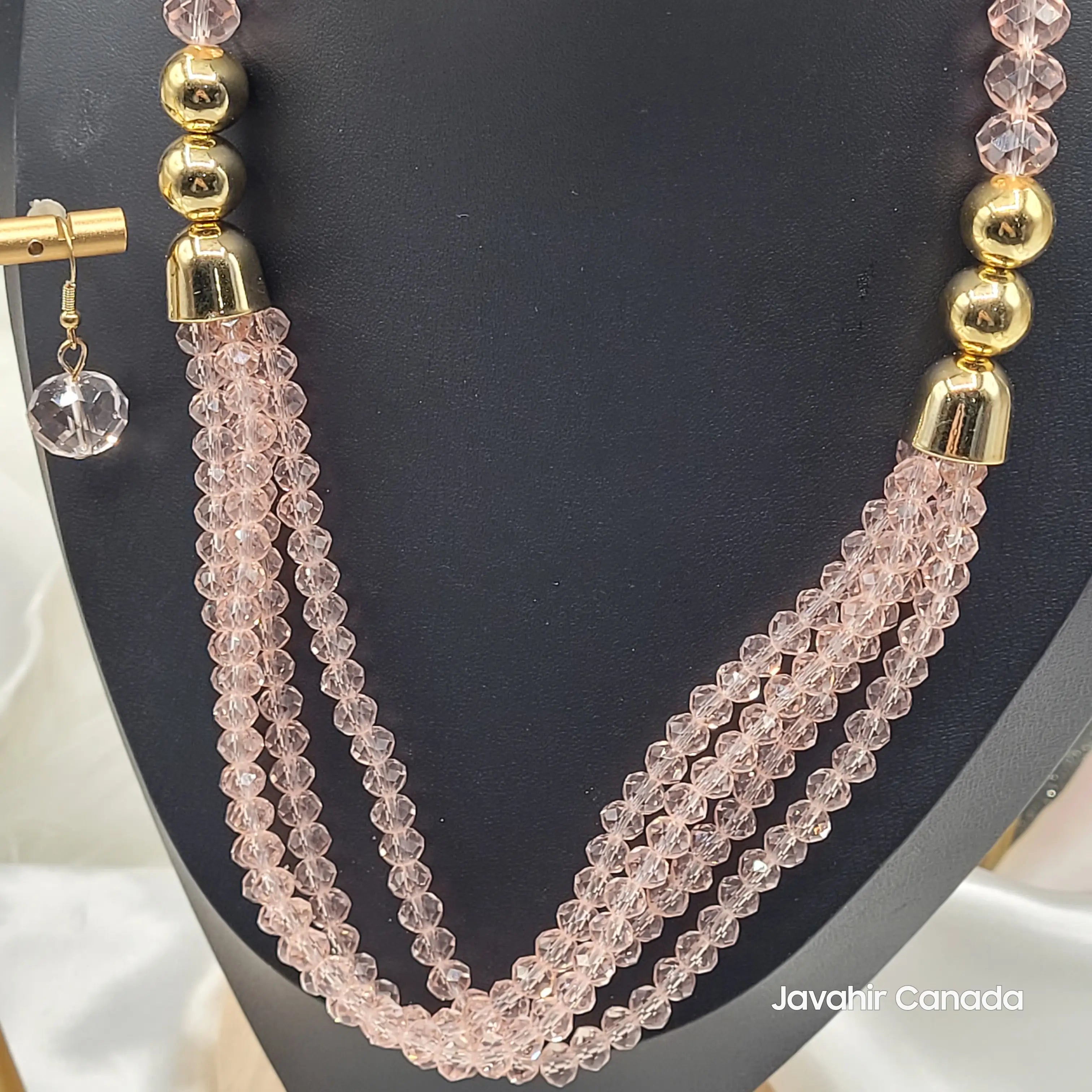 Close-up of the champagne pink JV161 necklace showing the twisted triple-strand crystal beading and glossy gold-tone detailing. Matching pink earring on display stand.