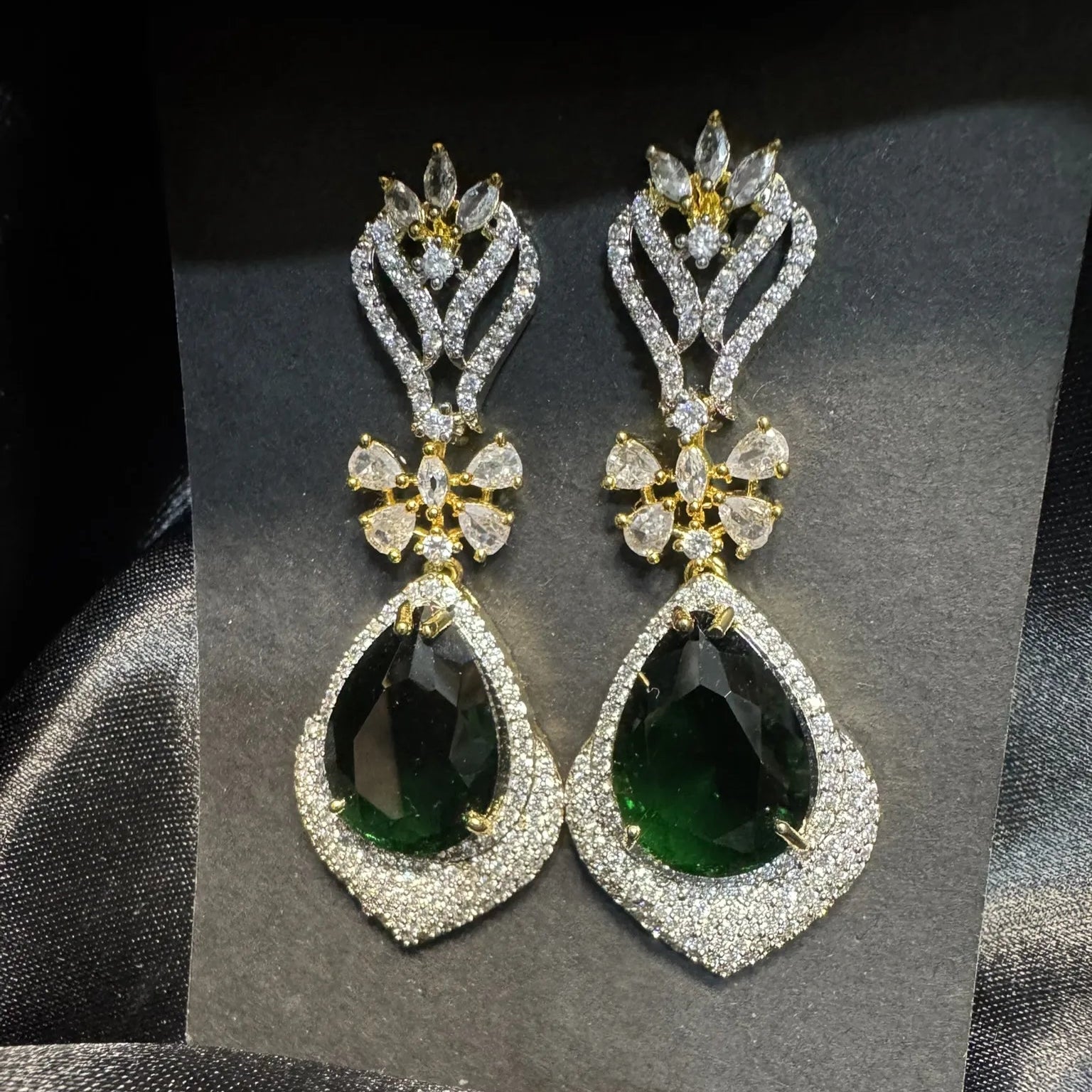 Pair of earrings with emerald gemstones and American diamonds stones on a gray card.