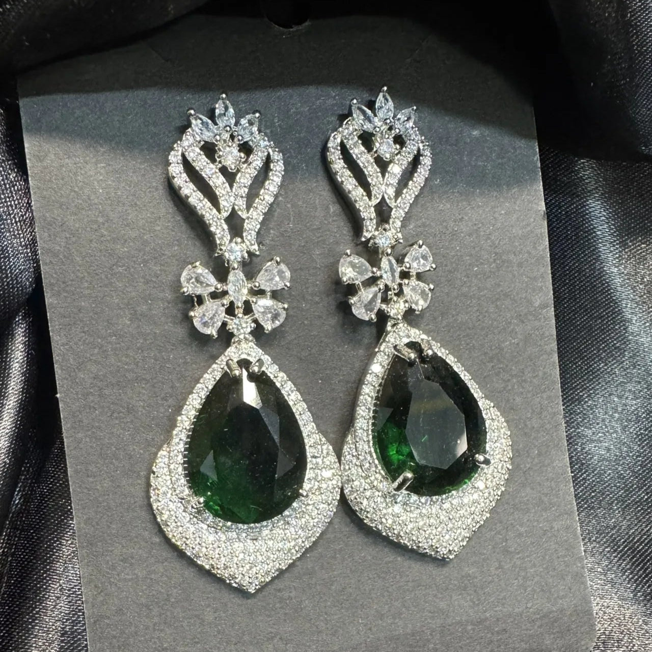 Pair of earrings with American diamonds and green gemstones on a gray card against a black satin background