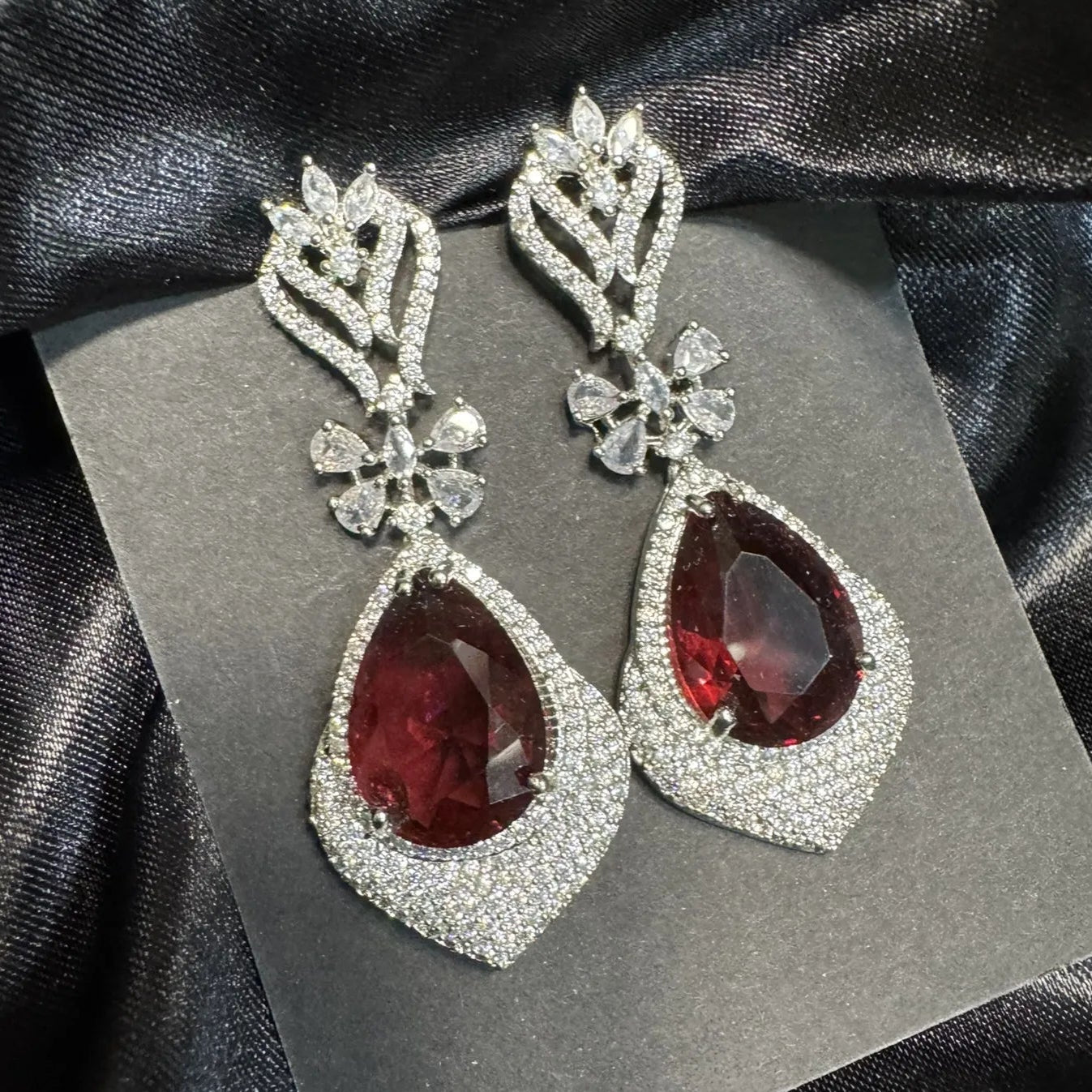 Pair of earrings with American diamonds and maroon gemstones and silver accents on a gray card.