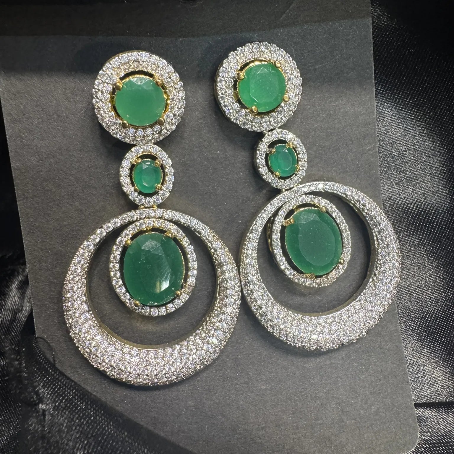 Pair of earrings with green gemstones and silver circular designs with American diamonds on a black background