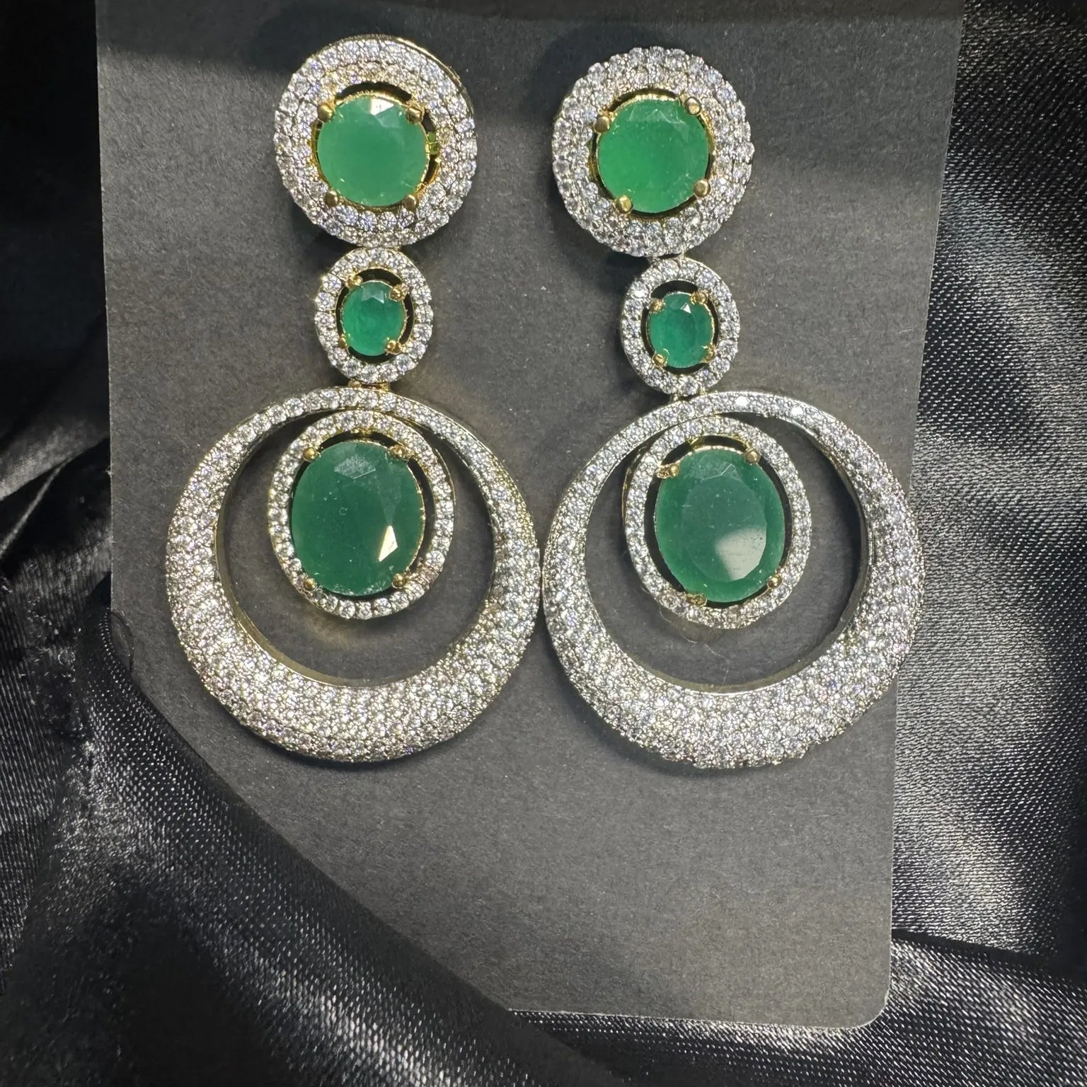 Pair of earrings with green gemstones and silver American diamonds accents on a black satin background