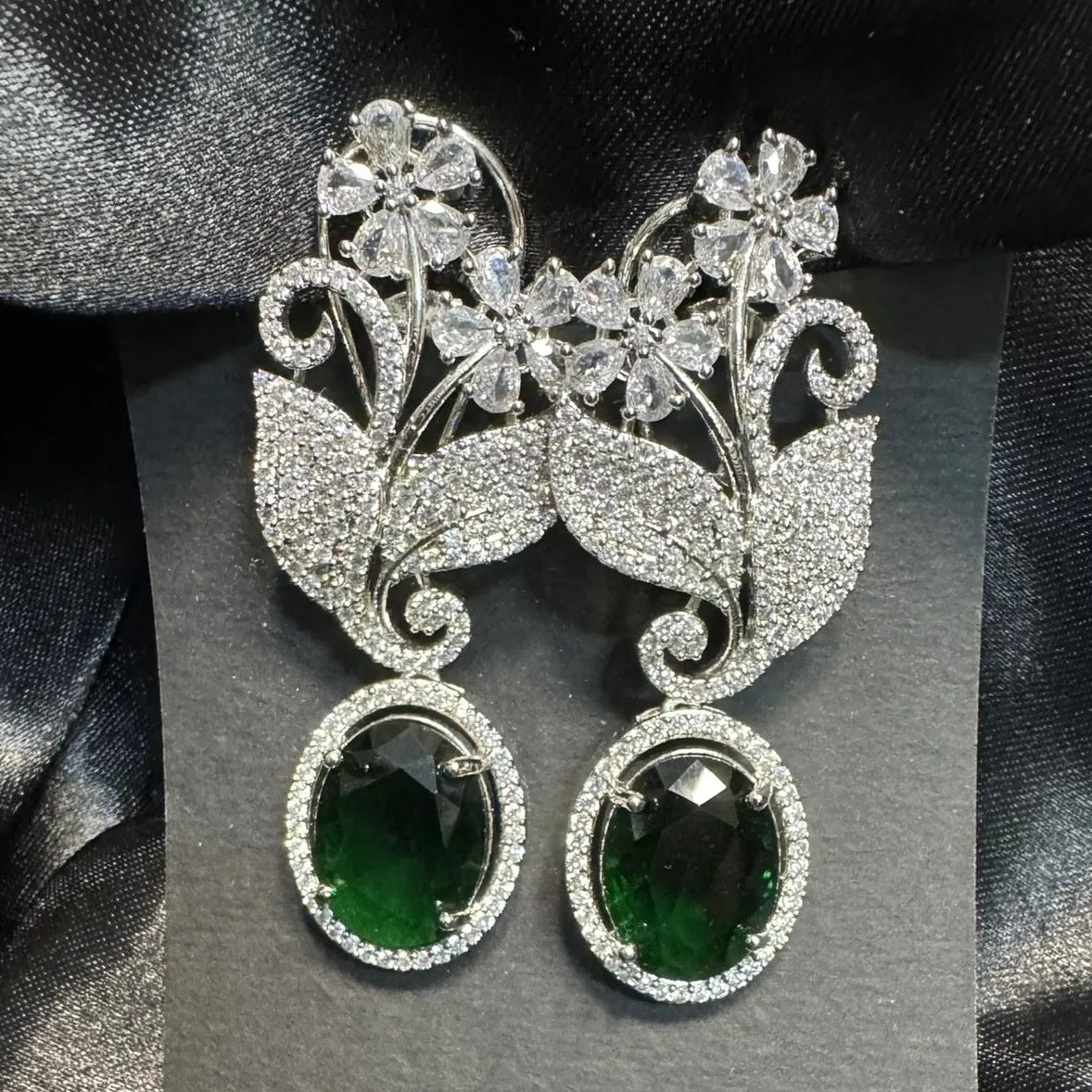 Ornate earrings with green gemstones on a gray card against a satin background