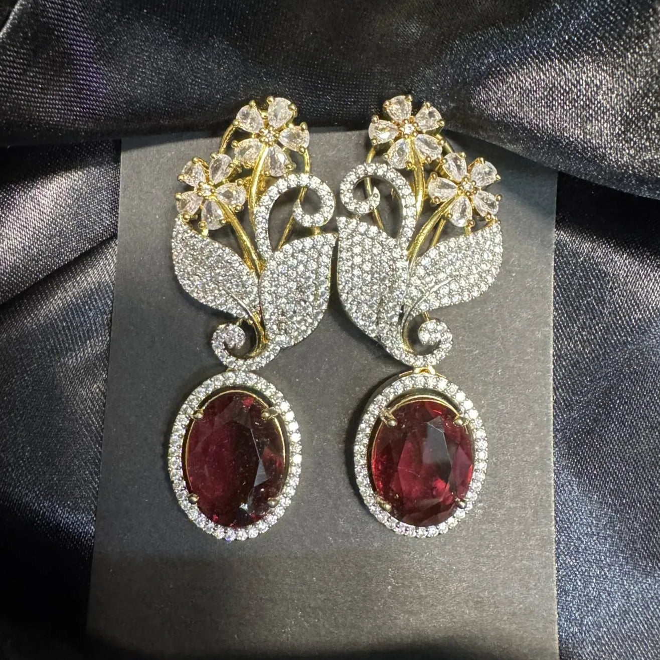 Pair of earrings with red gemstones and floral designs on a gray card against a dark fabric background.