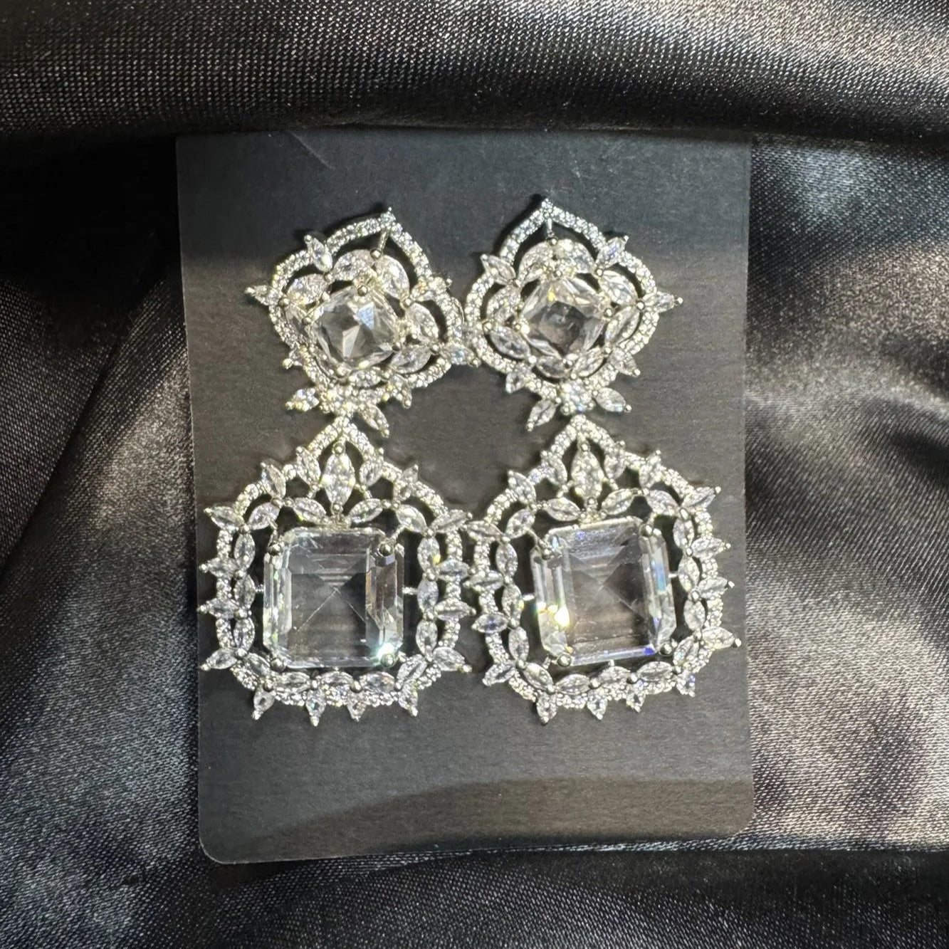 Pair of ornate earrings with clear gemstones on a black card against a black fabric background