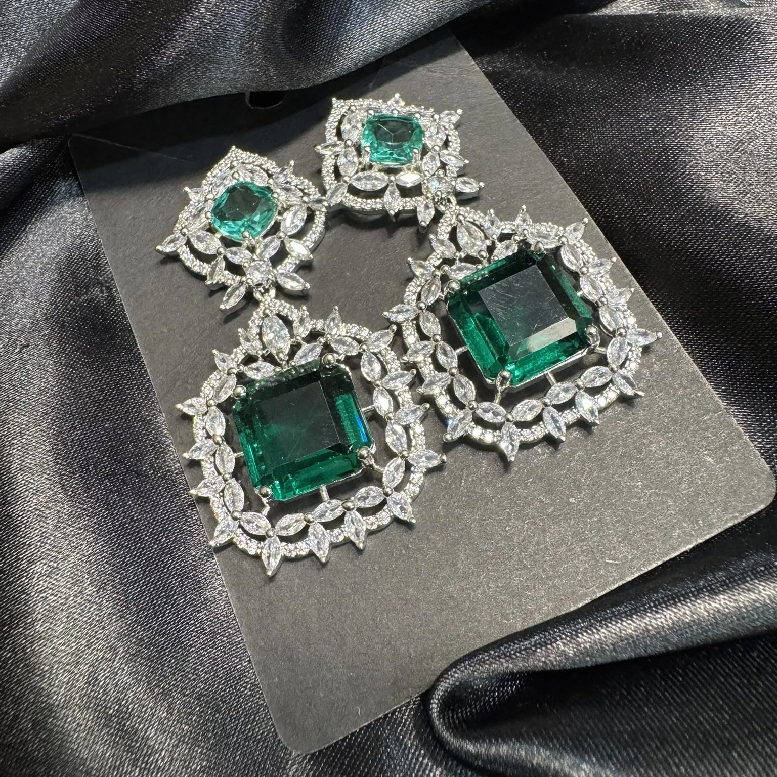 Pair of emerald green gemstone earrings on a gray card with black satin fabric background
