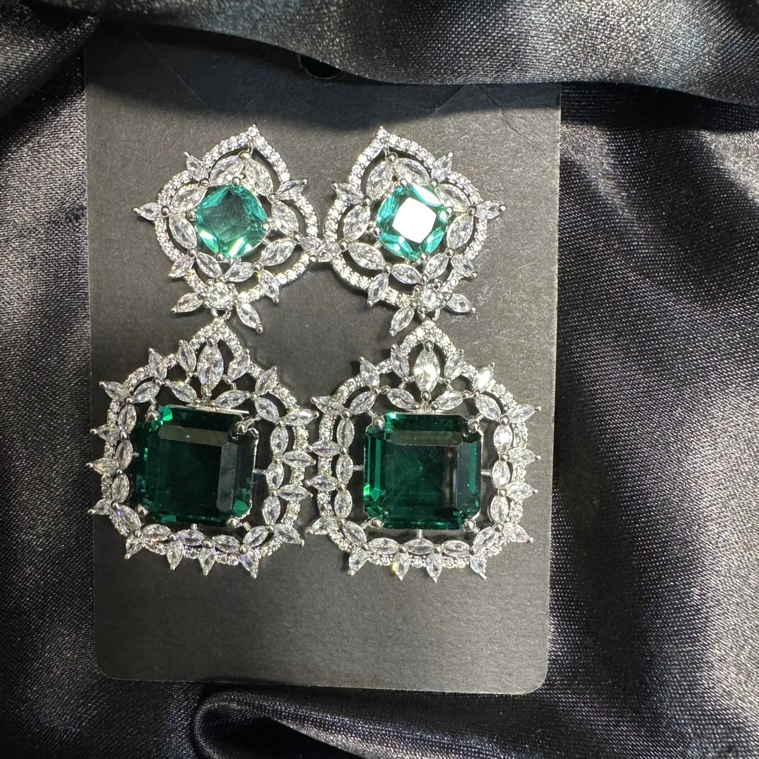 Pair of emerald green earrings with diamond-like stones on a gray card against a black satin background.